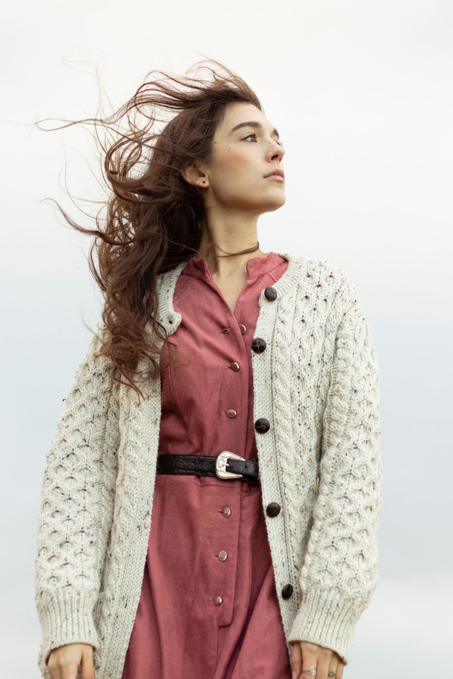 Wind in hair in a cream cardigan and rose dress against a pale sky